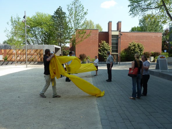 Eine Gruppe von Menschen läuft mit einer 15m langer gelben Fahne über den Brunnenplatz. Im Hintergrund ist der Gemeinschaftspavillon und die Katholische Kirche zu sehen.