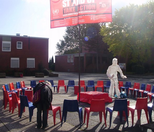 Ein Stuhlkreis aus roten und blauen Bofinger Stühlen auf der Mitte des Brunnenplatzes. In der Mitte steht eine Maschendraht-Figur eingewickelt in transparenter Folie (Der gläserne HuMu).
