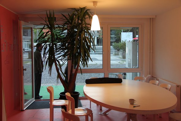 Ein großer weißer Tisch und Stühlen in der Mitte eines Raumes. Im Hintergrund steht eine große Palmenpflanze vor einer Fensterwand, durch die man den Brunnenplatz erkennen kann.