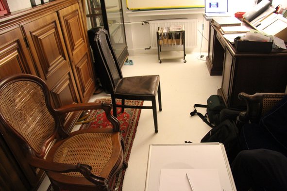 Ein Büroraum. Links ein Schrank aus dunklem Holz, davor stehen zwei Stühle. Rechts ein Schreibtisch mit Unterlagen.