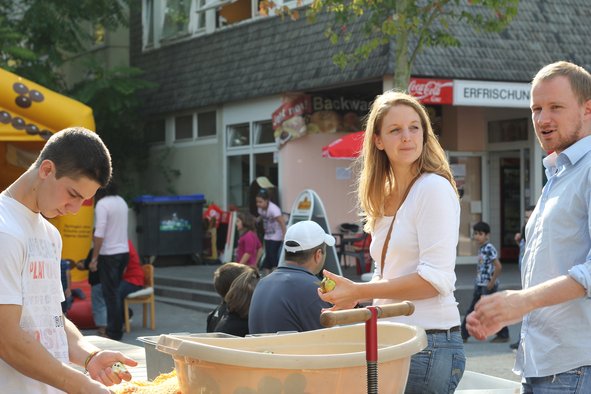 Jessica Schlierenkamp, Christian Leye und Daniel Matzkevitch stehen auf dem Brunnenplatz um eine Plastikwanne gefüllt mit Äpfeln. Sie sind ins Gespräch vertieft und zerkleinern die Äpfel.
