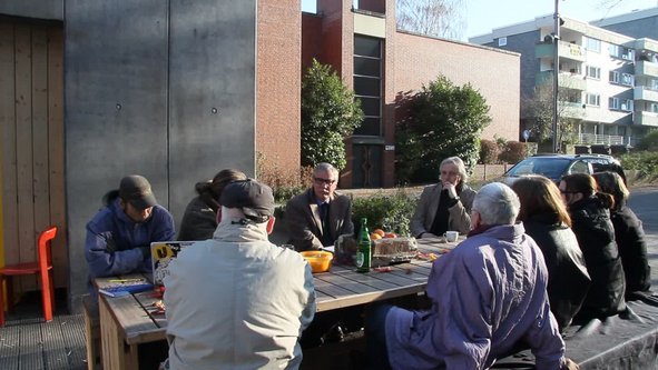 Eine Gruppe von Menschen sitzt an einem der Tische des Gemeinschaftspavillons Brunnenplatz 1. Stadtbaurat Ernst Kratsch sitzt der Kamera zugewandt.